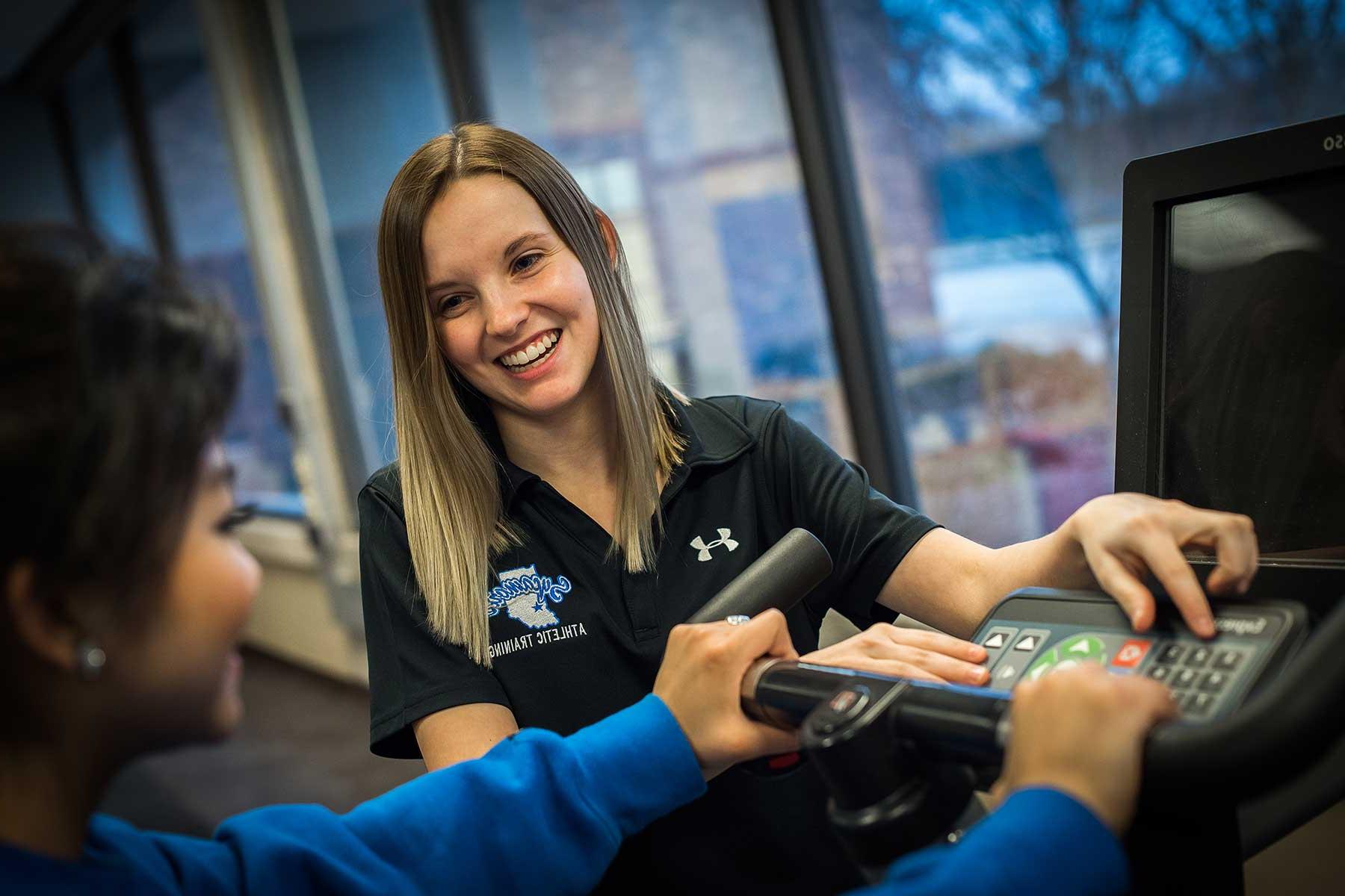 A SMILING FEMALE STUDENT IN A BLUE SWEATER HOLDS ONTO THE HANDLEBARS OF A STATIONARY BIKE WHILE ANOTHER SMILING, FEMALE STUDENT IN A BLACK SYCAMORES ATHLETIC TRAINING POLO SHIRT PUSHES BUTTONS ON THE KEYPAD. A LARGE WINDOW IS VISIBLE BEHIND THEM.