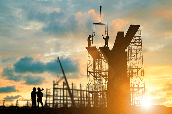 The frame and scaffolding of a building under construction is backlit by the rising sun. Two workers at the very top lift the arms to steady a section of frame being lowered to them by a construction crane (not visible), while two workers on the ground talk and one motions toward the building frame with his hand. All workers are in shadow but can be seen to wear hardhats.