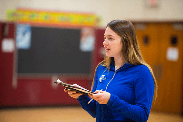 A woman with dark brown hair and blonde highlights stands in a gymnasium and holds a clipboard. She looks off into the distance. She is wearing a long-sleeved, blue ISU quarter-zip. Doors, a maroon-colored wall, and a large black bulletin board with a colorful border on the top are blurred but visible in the background.